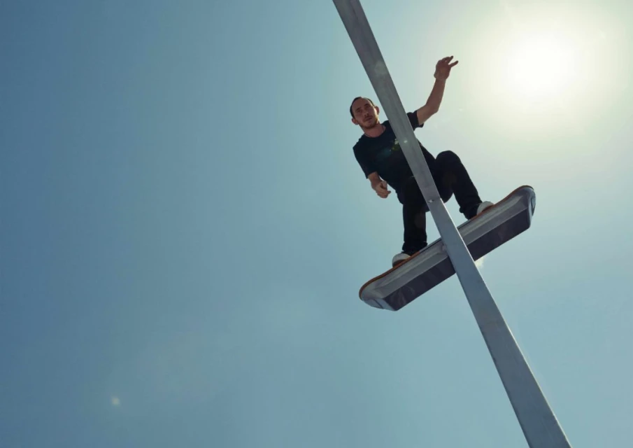 A low-angle shot captures a male skateboarder balancing on a street light, set against a clear blue sky with the sun shining brightly.