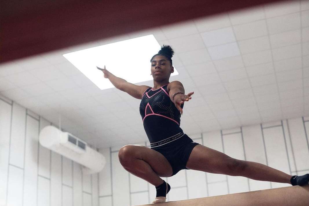 An African American woman gymnast in a black leotard and athletic shorts performs a balance beam routine in a bright indoor space.