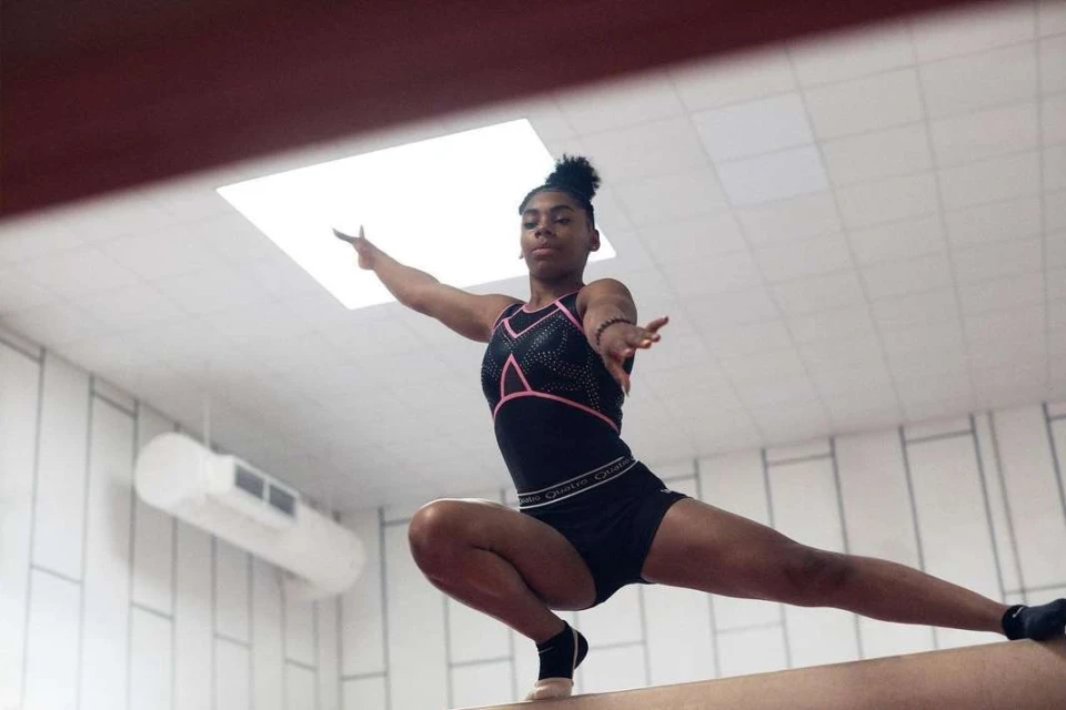 An African American woman gymnast in a black leotard and athletic shorts performs a balance beam routine in a bright indoor space.