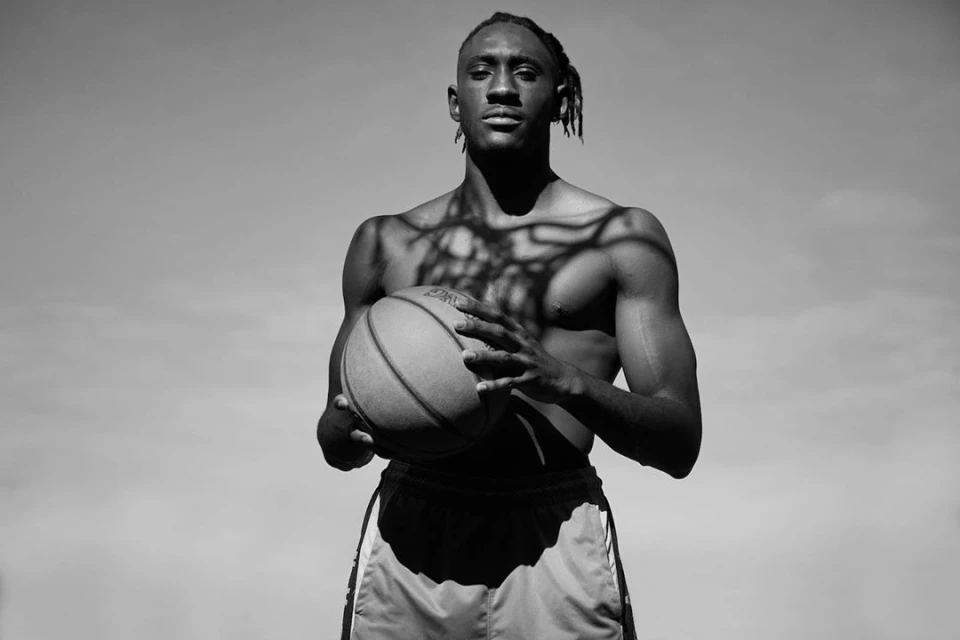 Black and white portrait of shirtless basketball player with dreadlocks holding ball against light sky, tree shadows falling on torso.