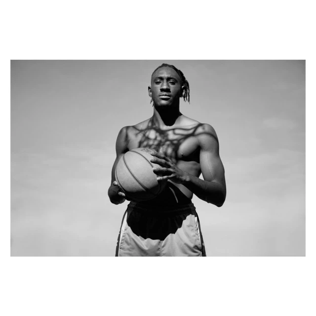 Striking black and white portrait of athletic man with dreadlocks holding basketball outdoors with tree shadows on his torso.