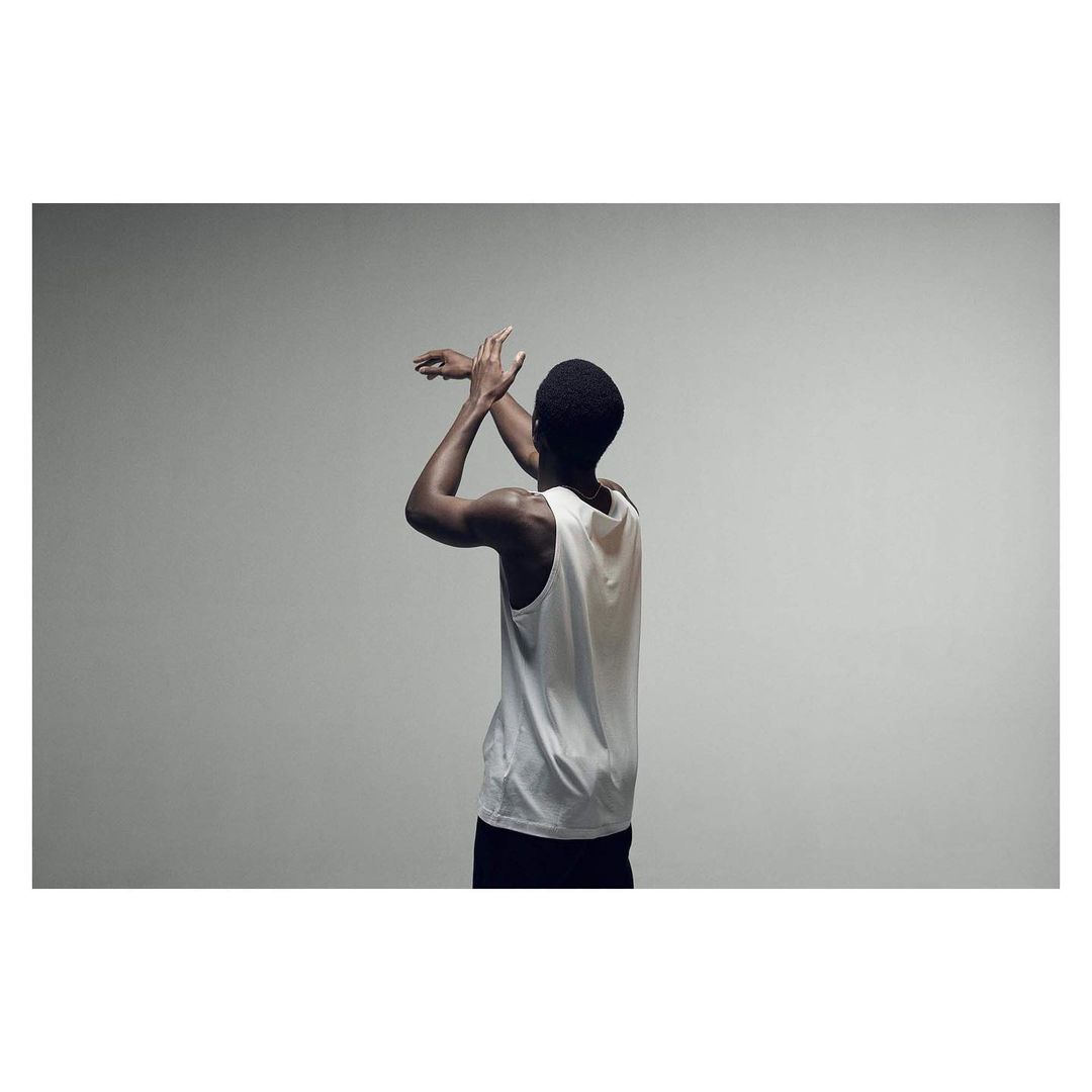Rear view of a Black man with short hair, wearing a white tank top and black shorts, with arms raised indoors against a gray backdrop.