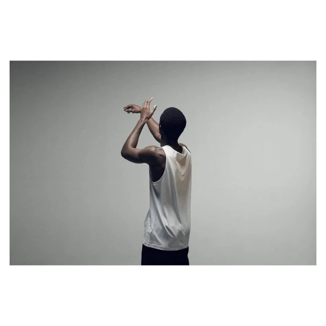 Rear view of a Black man with short hair, wearing a white tank top and black shorts, with arms raised indoors against a gray backdrop.