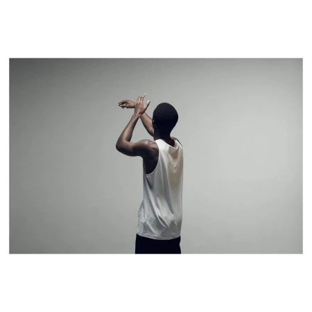 Rear view of a Black man with short hair, wearing a white tank top and black shorts, with arms raised indoors against a gray backdrop.