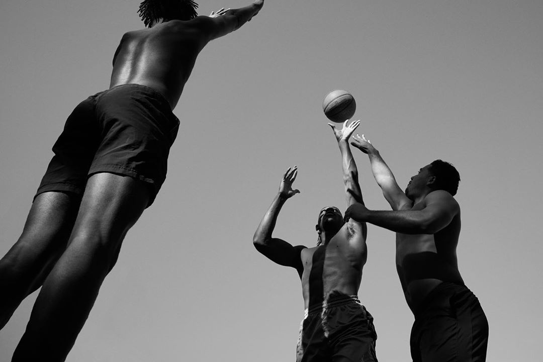 Three African American men playing basketball outdoors in monochrome. One man jumps for the ball, while the other two reach up toward it.