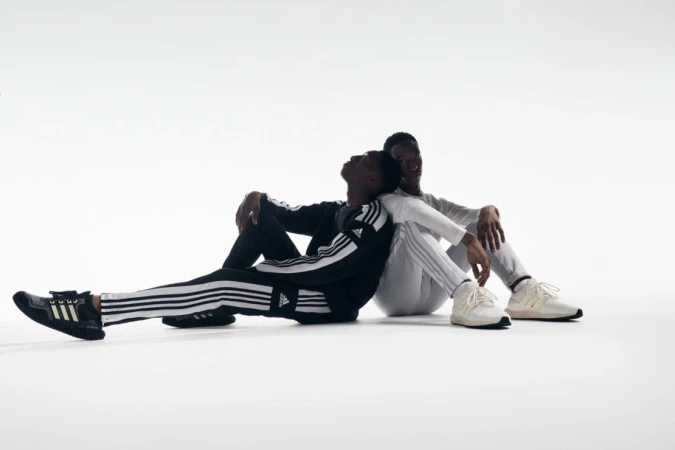 Two Black adults in Adidas tracksuits sit back-to-back against a white background in a studio, showcasing athletic apparel and footwear.