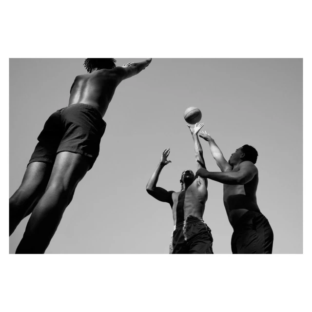 Monochrome photo of three athletic men playing basketball under a bright sky, one leaping for the ball, captured from a low angle.