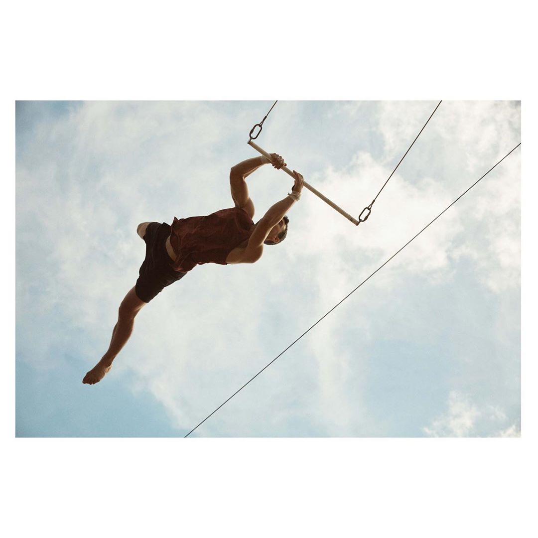 A man is suspended mid-air on a trapeze bar under a partly cloudy sky, performing acrobatics with outstretched arms and legs.