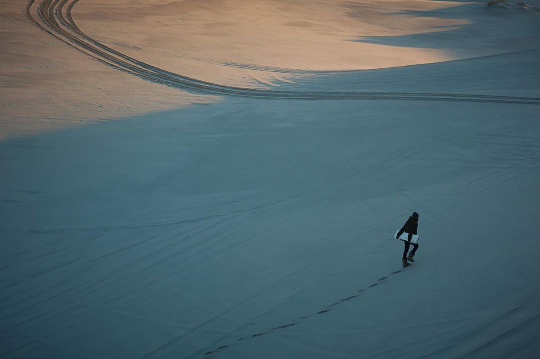 A lone figure walks across a vast, snow-covered landscape under a soft, muted sky, leaving a trail of footprints in the snow.