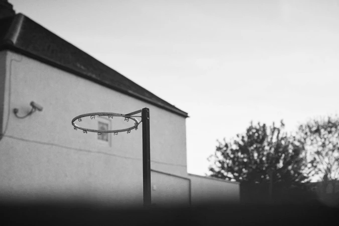 Monochrome image of a weathered basketball hoop and pole standing outdoors near a building and trees against a bright sky.