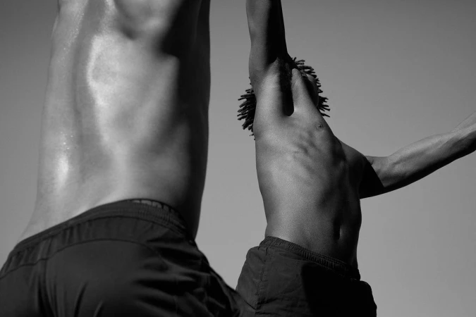 Black and white torso shot of two athletic men with dreadlocks outside in sportswear against a plain sky background during daylight hours.