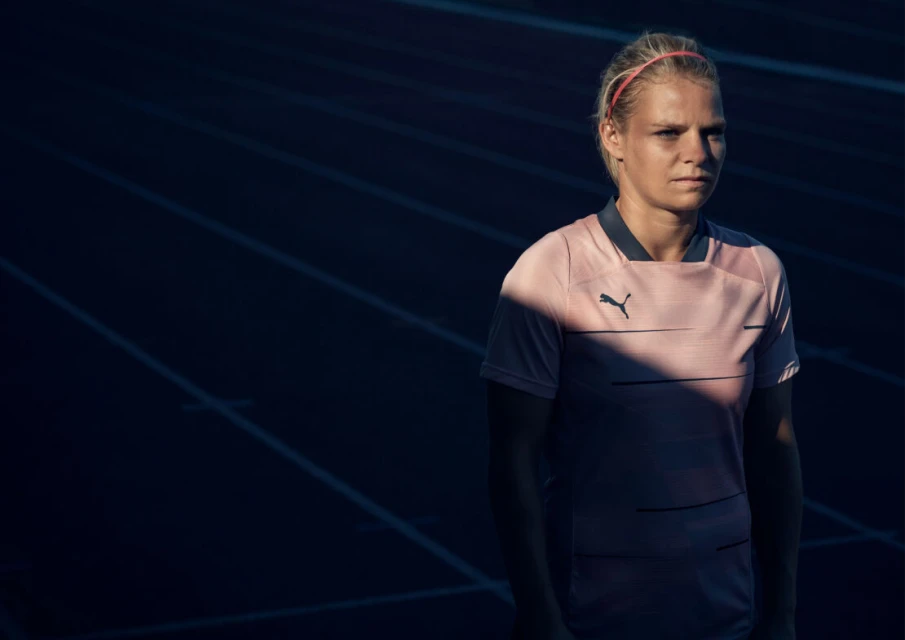 Female athlete in pink Puma shirt stands on running track under dramatic lighting, showcasing fitness and sportswear fashion.