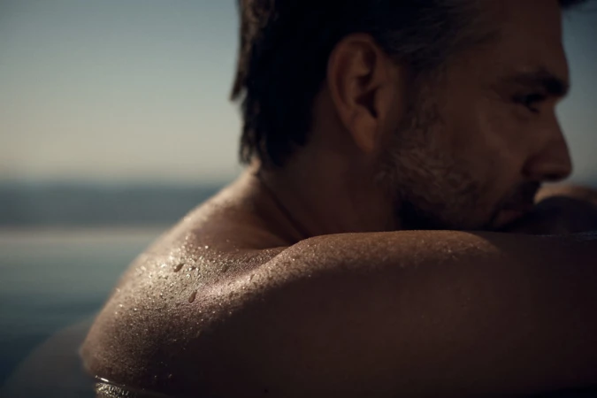Close-up profile of a shirtless man with water droplets on his shoulders, captured outdoors near a body of water at dusk by Olly Burn.