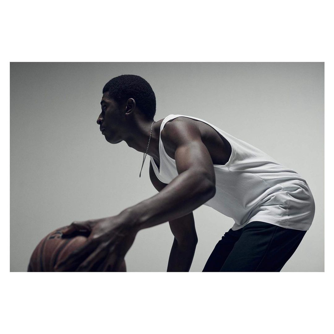 Athletic African-American man dribbling basketball indoors, wearing a white tank top and dark shorts, lit by studio lighting.