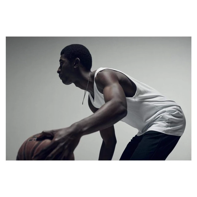 Athletic African-American man dribbling basketball indoors, wearing a white tank top and dark shorts, lit by studio lighting.