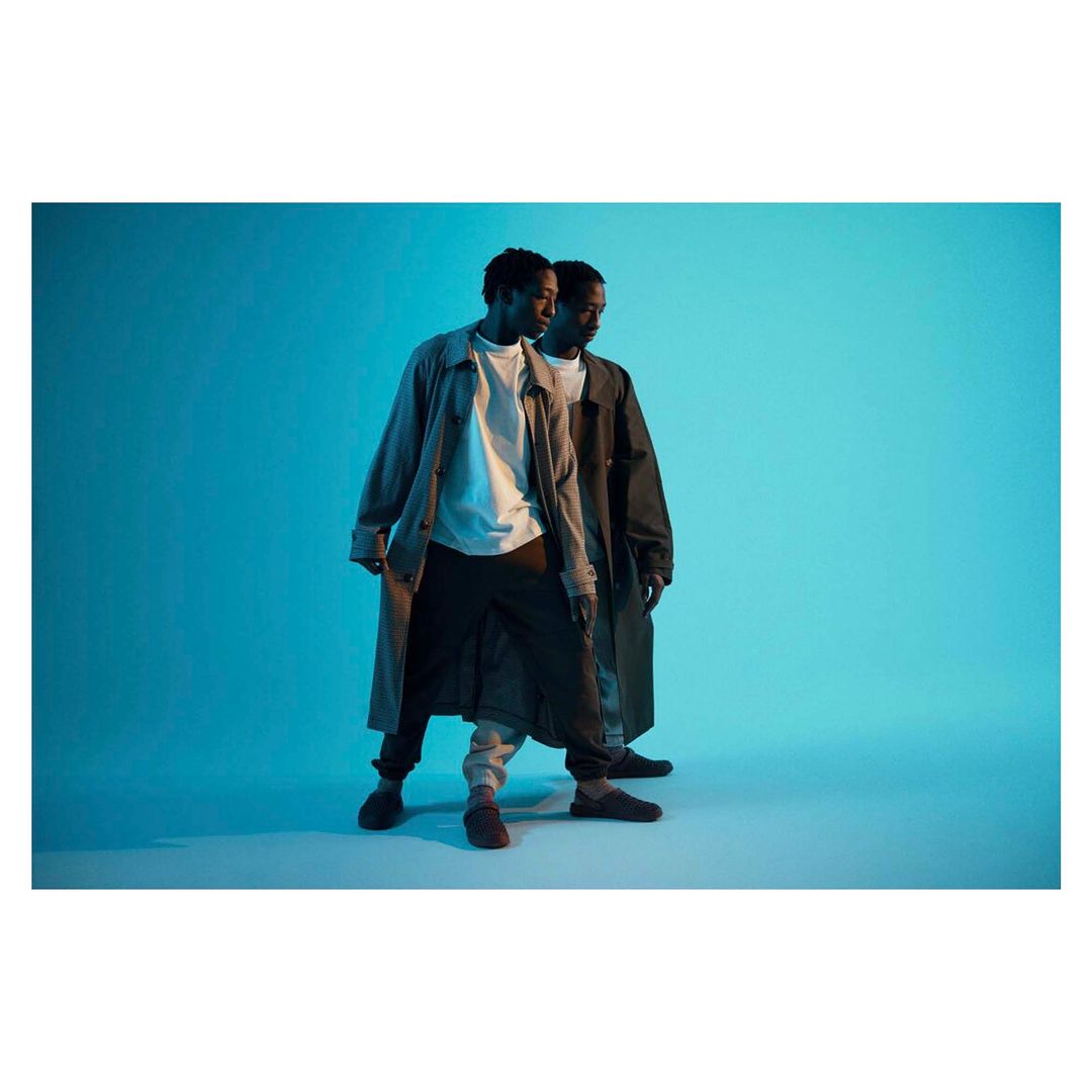 Two African American men in trench coats pose in a photo studio against a blue background with neutral facial expressions and casual attire.