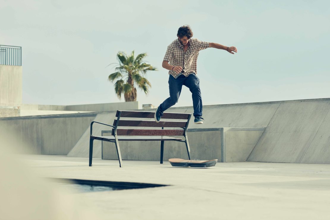 Male skateboarder jumping over bench in skatepark on sunny day with palm trees, wearing casual plaid shirt and jeans.
