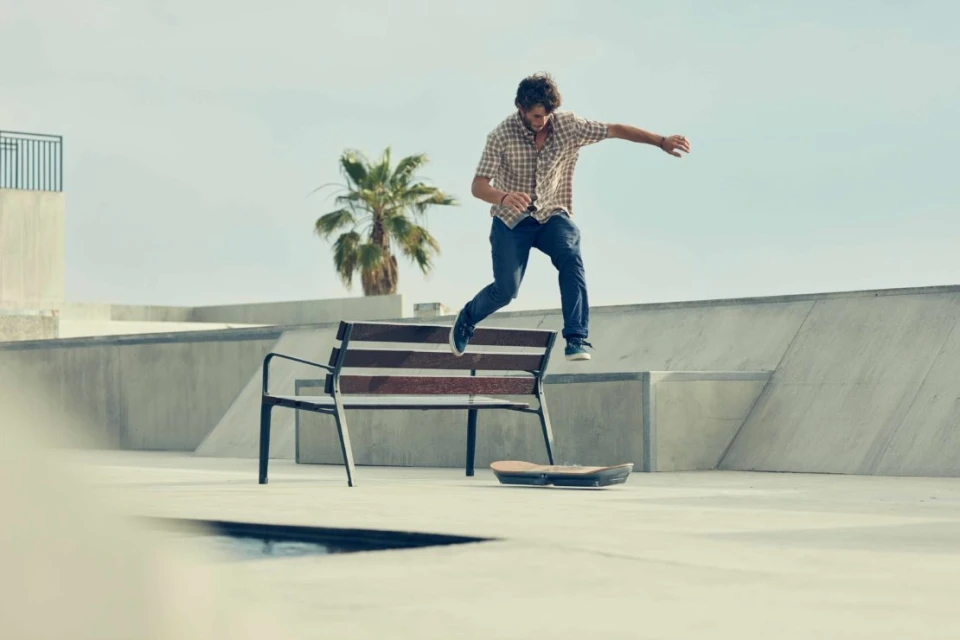 Male skateboarder jumping over bench in skatepark on sunny day with palm trees, wearing casual plaid shirt and jeans.