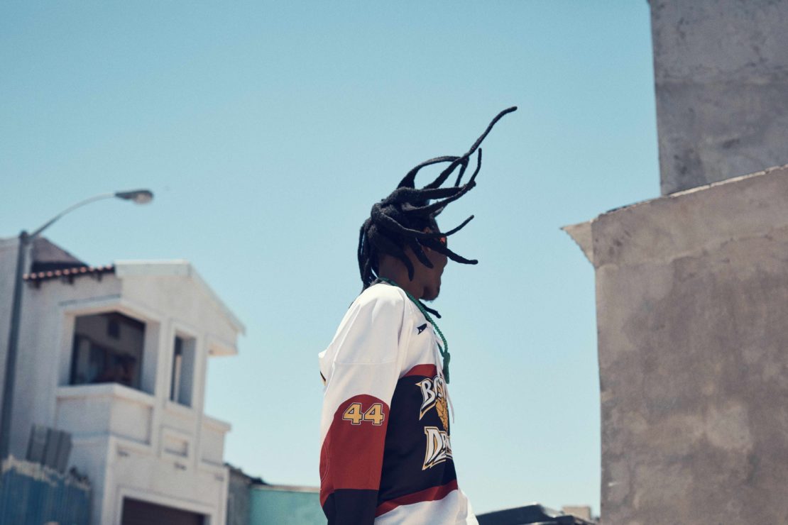 Man in hockey jersey outdoors against blue sky. Urban environment, street, and building visible in the background on a bright day.