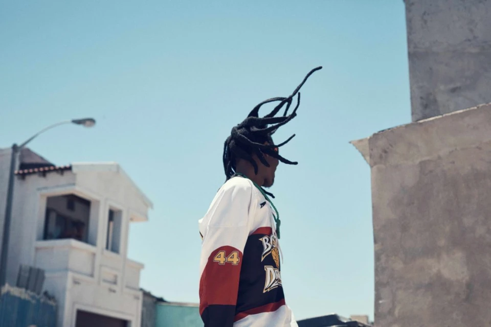 Man in hockey jersey outdoors against blue sky. Urban environment, street, and building visible in the background on a bright day.