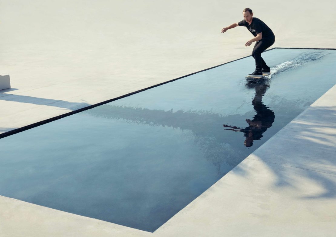 A man wearing black clothes is skateboarding on a pool of water that reflects his image on a bright sunny day in an urban outdoor environment.