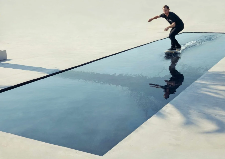 A man wearing black clothes is skateboarding on a pool of water that reflects his image on a bright sunny day in an urban outdoor environment.