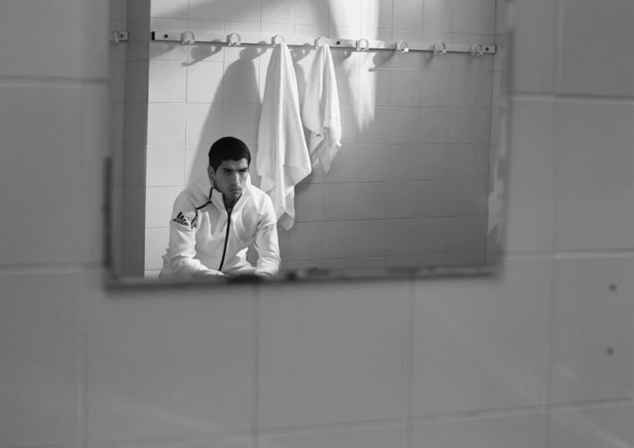 A man in a white jacket sits reflected in a mirror in a tiled room, with a towel rack and towels visible in the background.