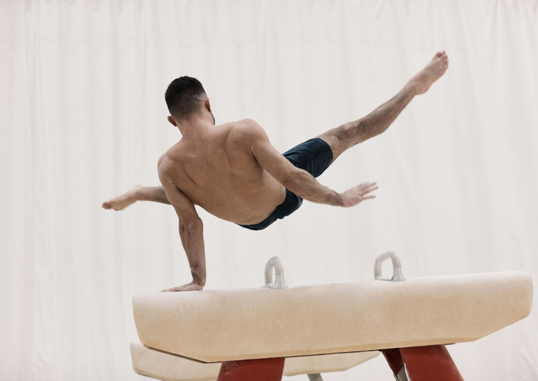A man performs gymnastics on a pommel horse inside a gymnasium, showcasing strength, flexibility, and balance against a blurred background.