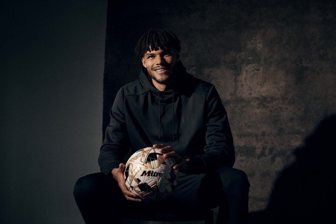 Portrait of a man with dark curly hair sitting and holding a soccer ball against a mottled grey background in a dimly lit indoor area.