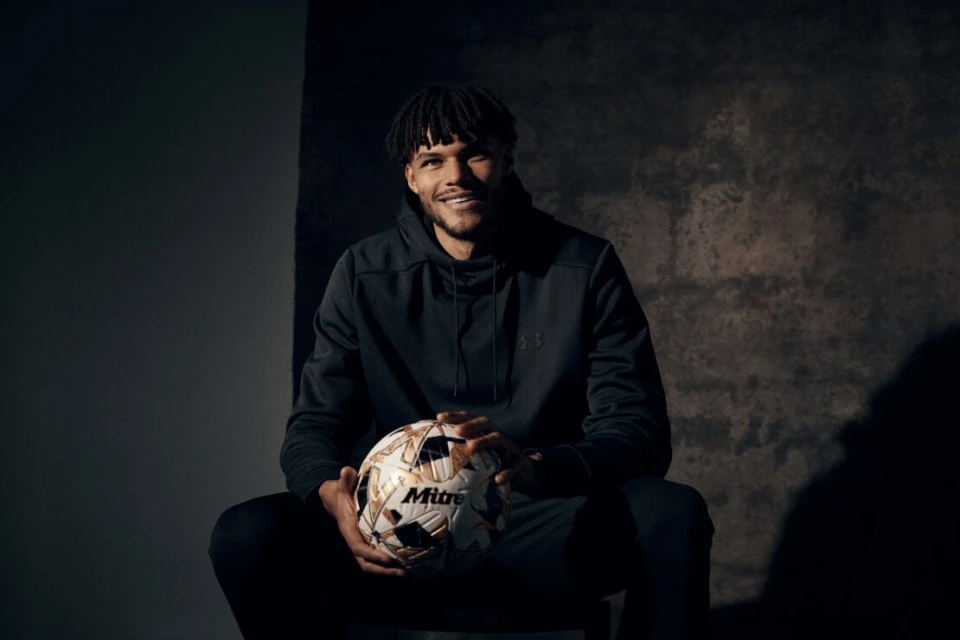 Portrait of a man with dark curly hair sitting and holding a soccer ball against a mottled grey background in a dimly lit indoor area.