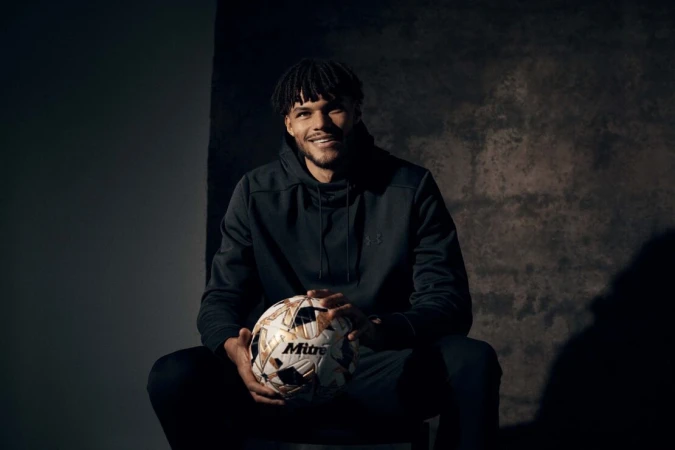 Portrait of a man with dark curly hair sitting and holding a soccer ball against a mottled grey background in a dimly lit indoor area.