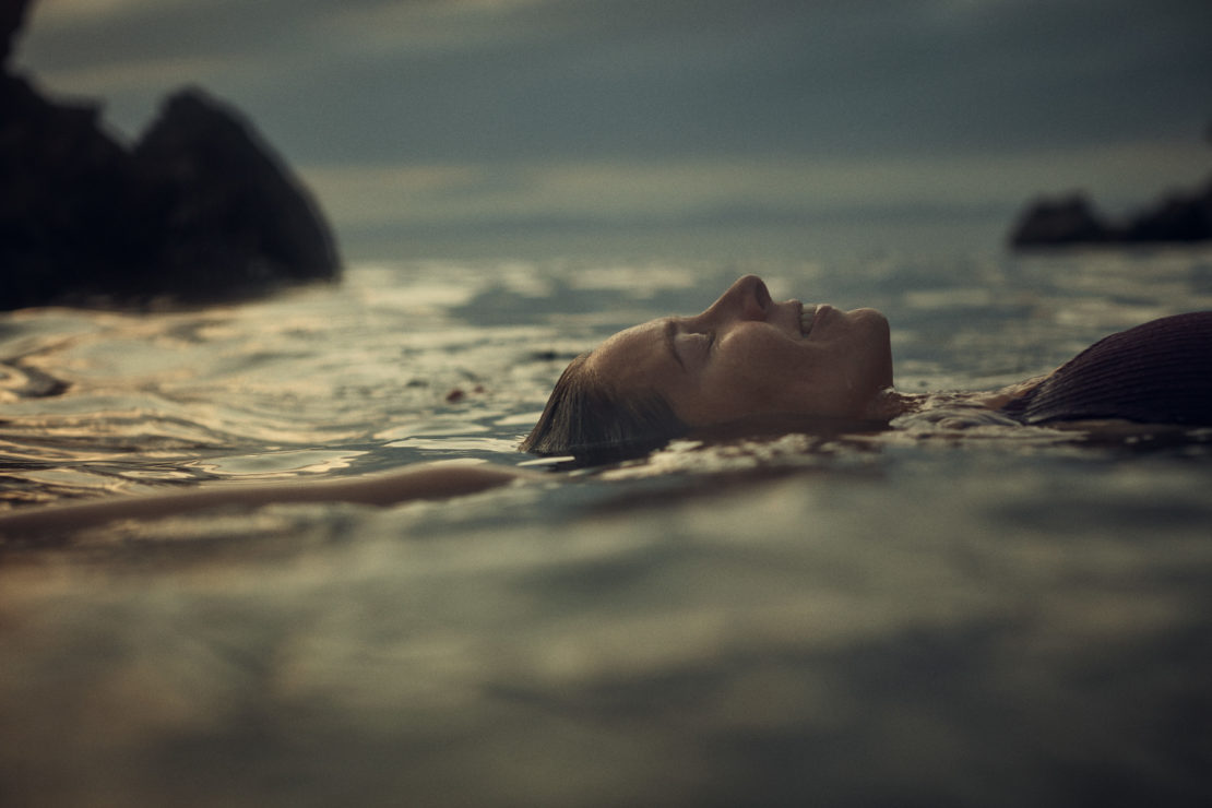 Profile of a person floating on their back in calm water with dark rocky formations visible in the distance under a dusky sky.