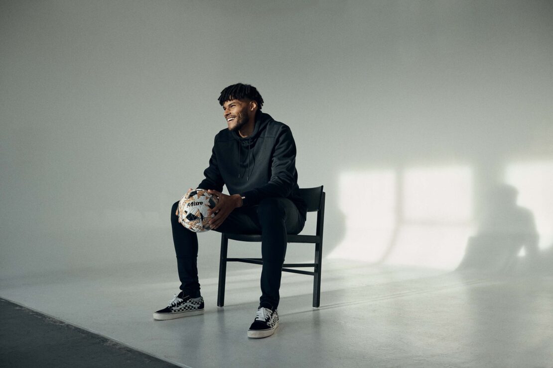 Adult male smiles while sitting on a black chair indoors, holding a soccer ball in his hands, and wearing a black hoodie and Vans sneakers.