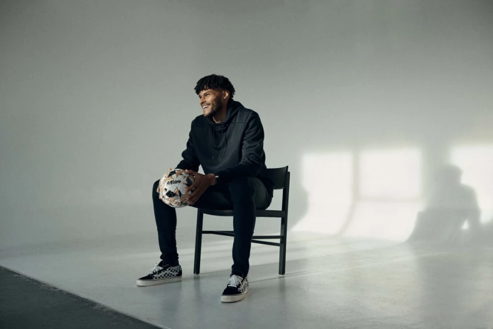 Adult male smiles while sitting on a black chair indoors, holding a soccer ball in his hands, and wearing a black hoodie and Vans sneakers.