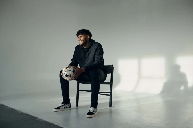 Adult male smiles while sitting on a black chair indoors, holding a soccer ball in his hands, and wearing a black hoodie and Vans sneakers.
