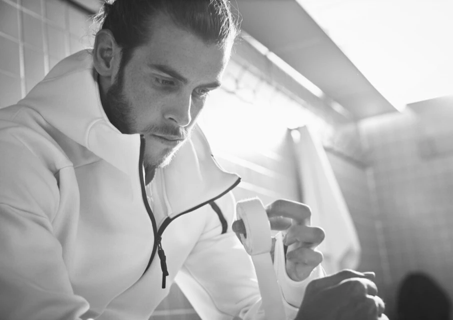Monochrome portrait of an adult man wrapping his hands, wearing a white hooded jacket in an indoor tiled environment, focusing on his hands.