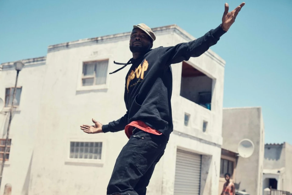 Adult male in a hoodie and cap posing outdoors against a backdrop of white buildings and a clear blue sky on a sunny day.