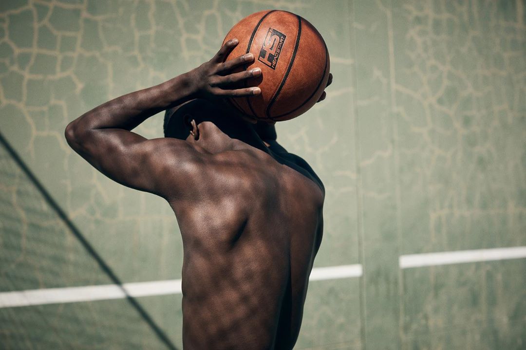 Athletic African American man holds a basketball behind his head, showcasing his muscular back against a textured wall.