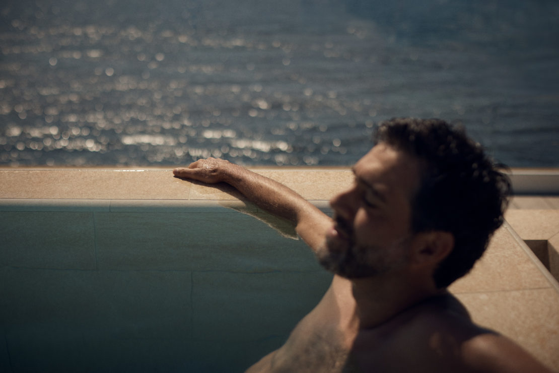 Man relaxing in a pool overlooking the ocean. Adult male with dark hair and a beard is partially submerged in the water.