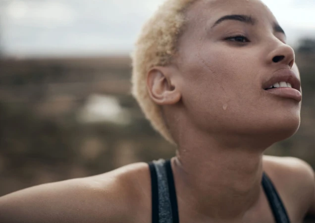 Close-up of a sweaty, fair-skinned woman with short blonde hair in athletic wear outdoors, looking upwards toward overcast sky.