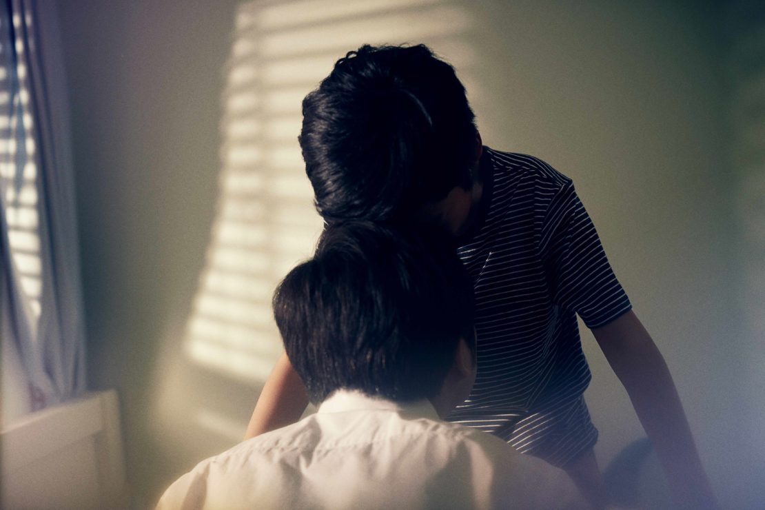 Two young boys, one in a striped shirt, standing in a room with light streaming through the window blinds, creating a cozy indoor scene.