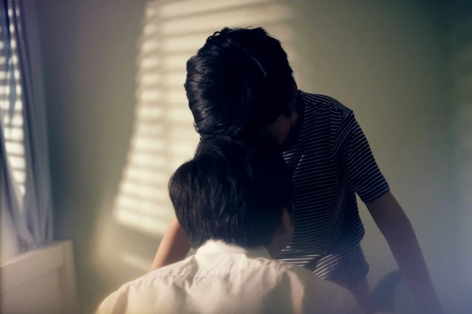 Two young boys, one in a striped shirt, standing in a room with light streaming through the window blinds, creating a cozy indoor scene.