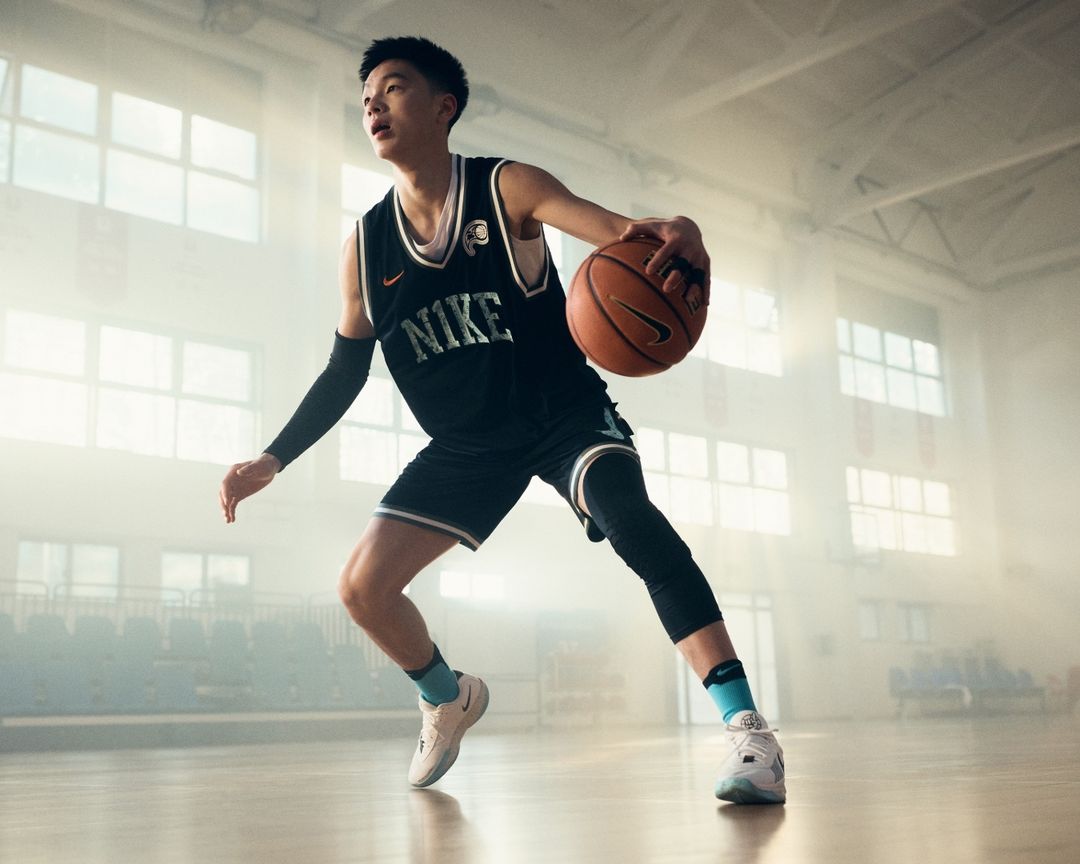 Male basketball player dribbling a ball in a gym, wearing a black Nike jersey, black shorts, knee pads, and white and gray sneakers.