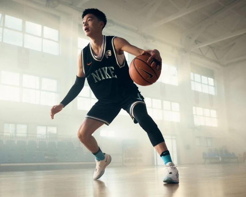 Male basketball player dribbling a ball in a gym, wearing a black Nike jersey, black shorts, knee pads, and white and gray sneakers.