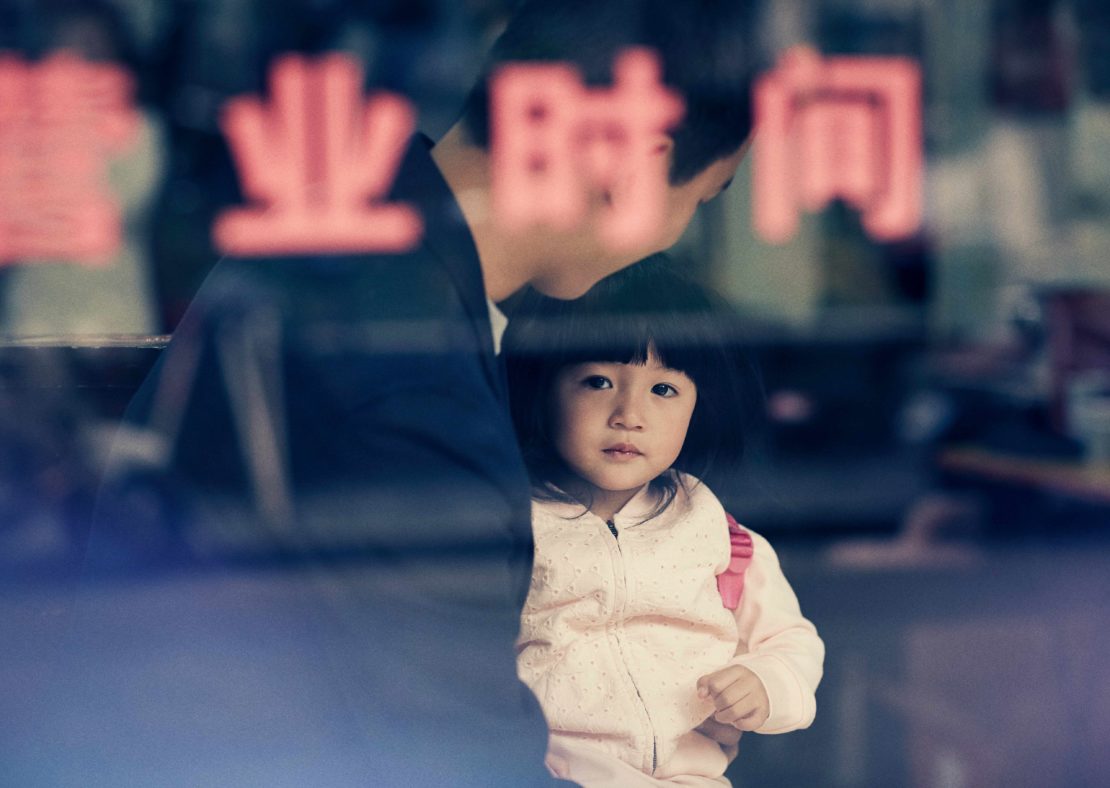 A young girl with dark hair looking at the camera, standing inside with a man behind a glass window with red Chinese text.