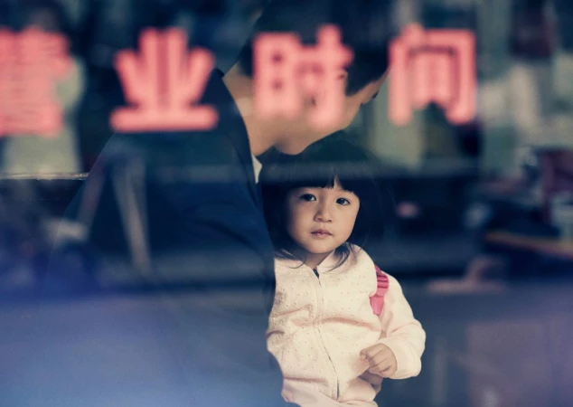 A young girl with dark hair looking at the camera, standing inside with a man behind a glass window with red Chinese text.