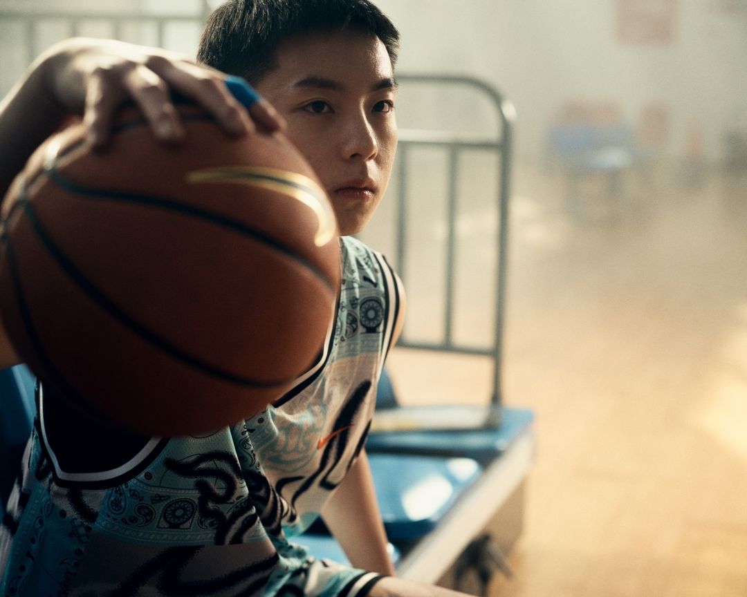 Young man in a sports jersey holds a basketball in a gymnasium, looking at the camera with a neutral expression.