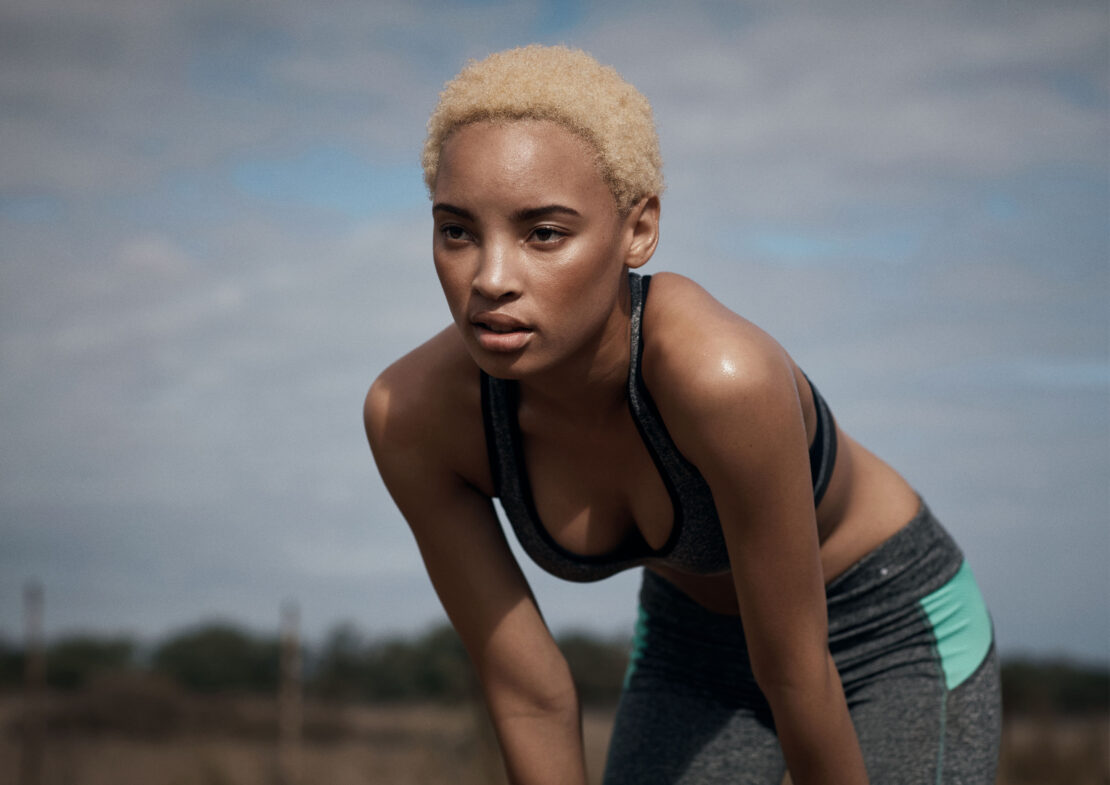 An athletic woman with short blonde hair rests outdoors in workout gear, leaning forward against a blurred background of sky and vegetation.
