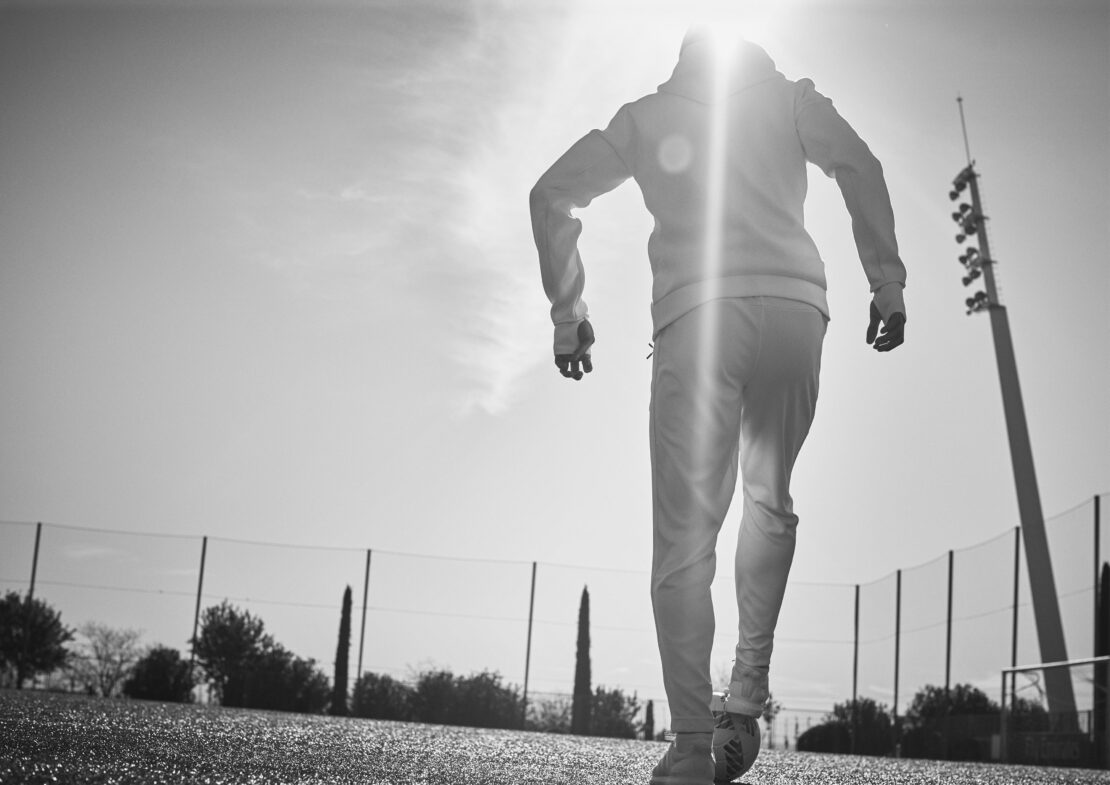 Black and white photo of an adult wearing a hooded sweatshirt walking on green artificial turf towards a tall light tower against the sky.