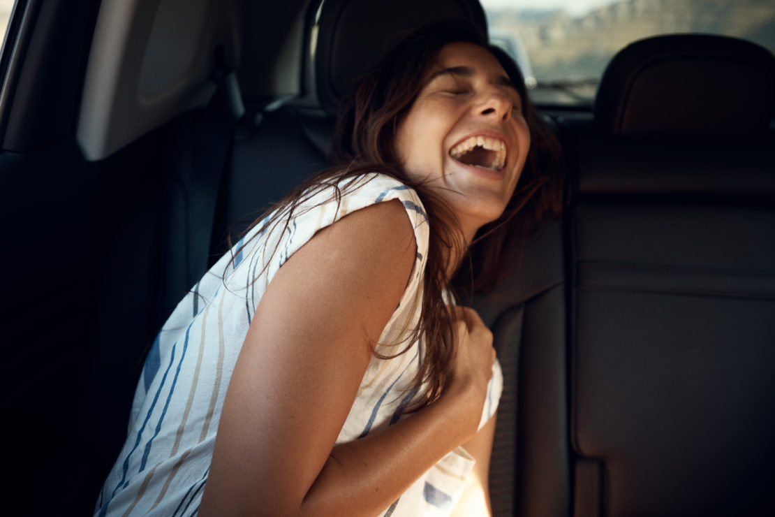 Brunette woman laughing in the backseat of a car, wearing a white and blue striped shirt, with a glimpse of an outdoor scene visible.